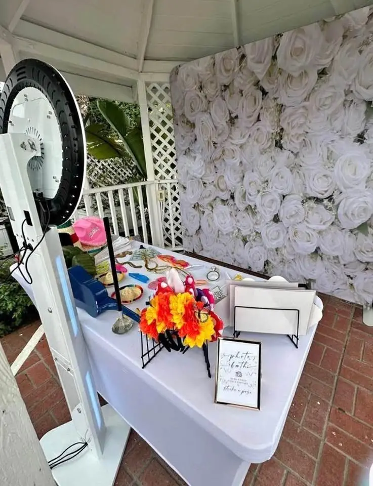 Open-air photo booth set up beside a white flower wall with a styled prop table